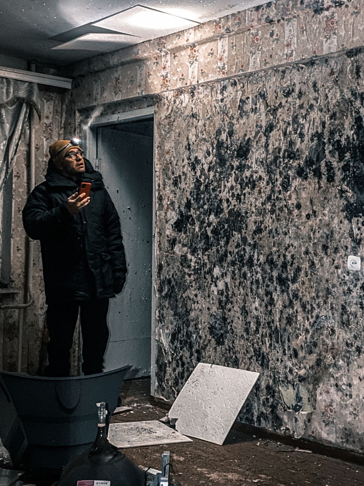 Person with a headlamp photographing the damaged interior of an old building with peeling wallpaper and fallen ceiling tiles